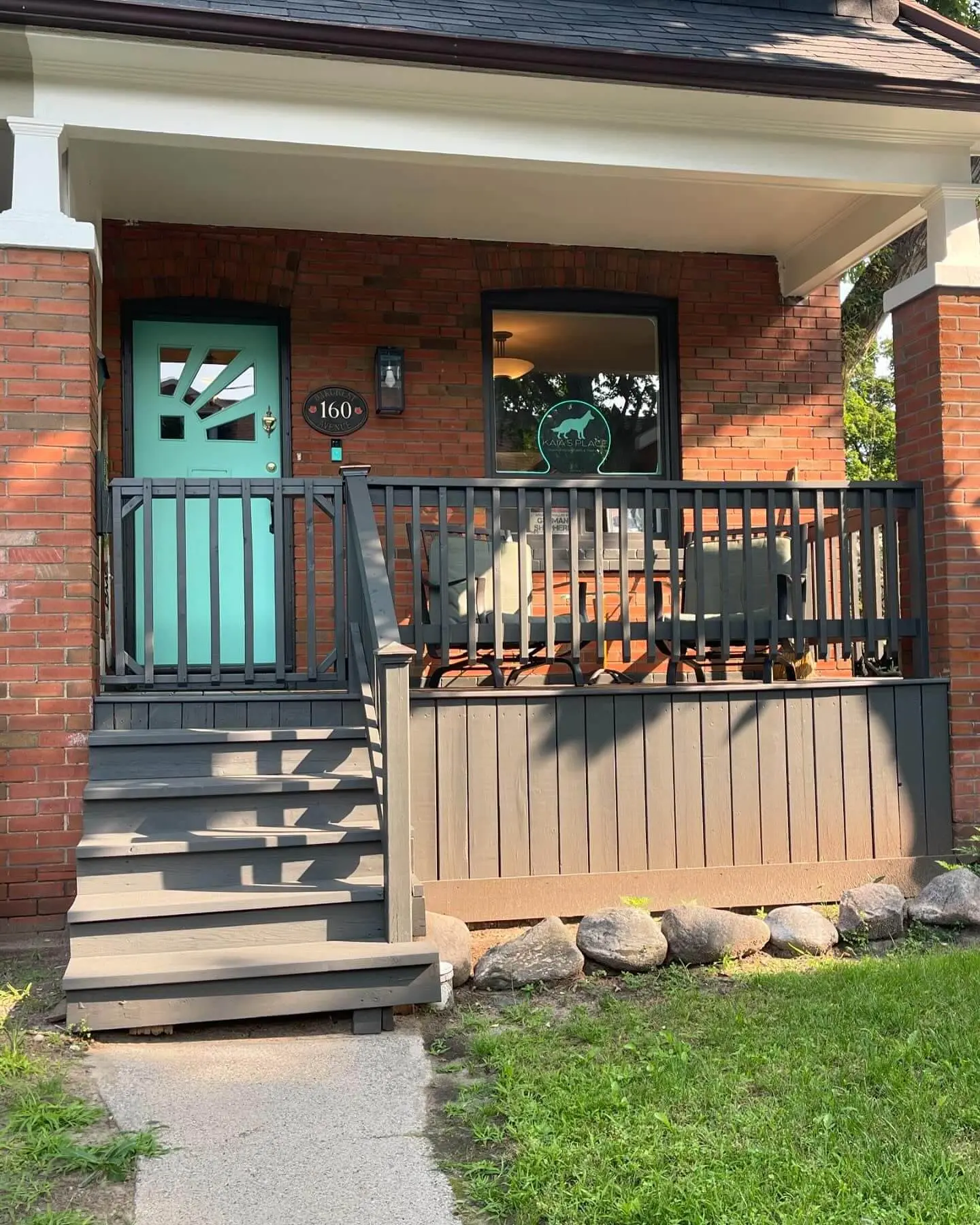 Cozy front porch of a red brick house with a turquoise front door, dark gray wooden steps, and railing, surrounded by greenery. Cozy front porch of a red brick house with a turquoise front door, dark gray wooden steps, and railing, surrounded by greenery.