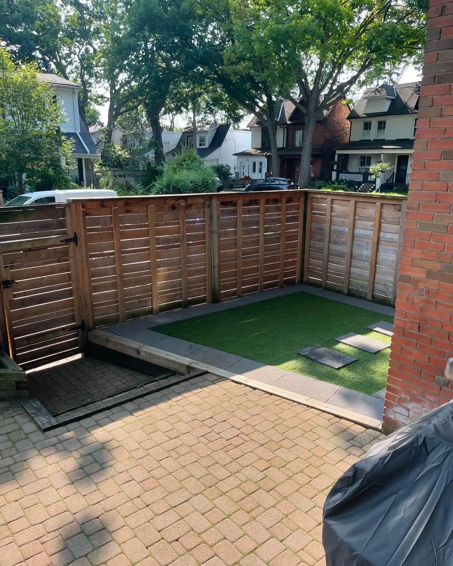View of a fenced backyard with artificial turf, brick wall, and wooden privacy fence, under the shade of large trees. View of a fenced backyard with artificial turf, brick wall, and wooden privacy fence, under the shade of large trees.