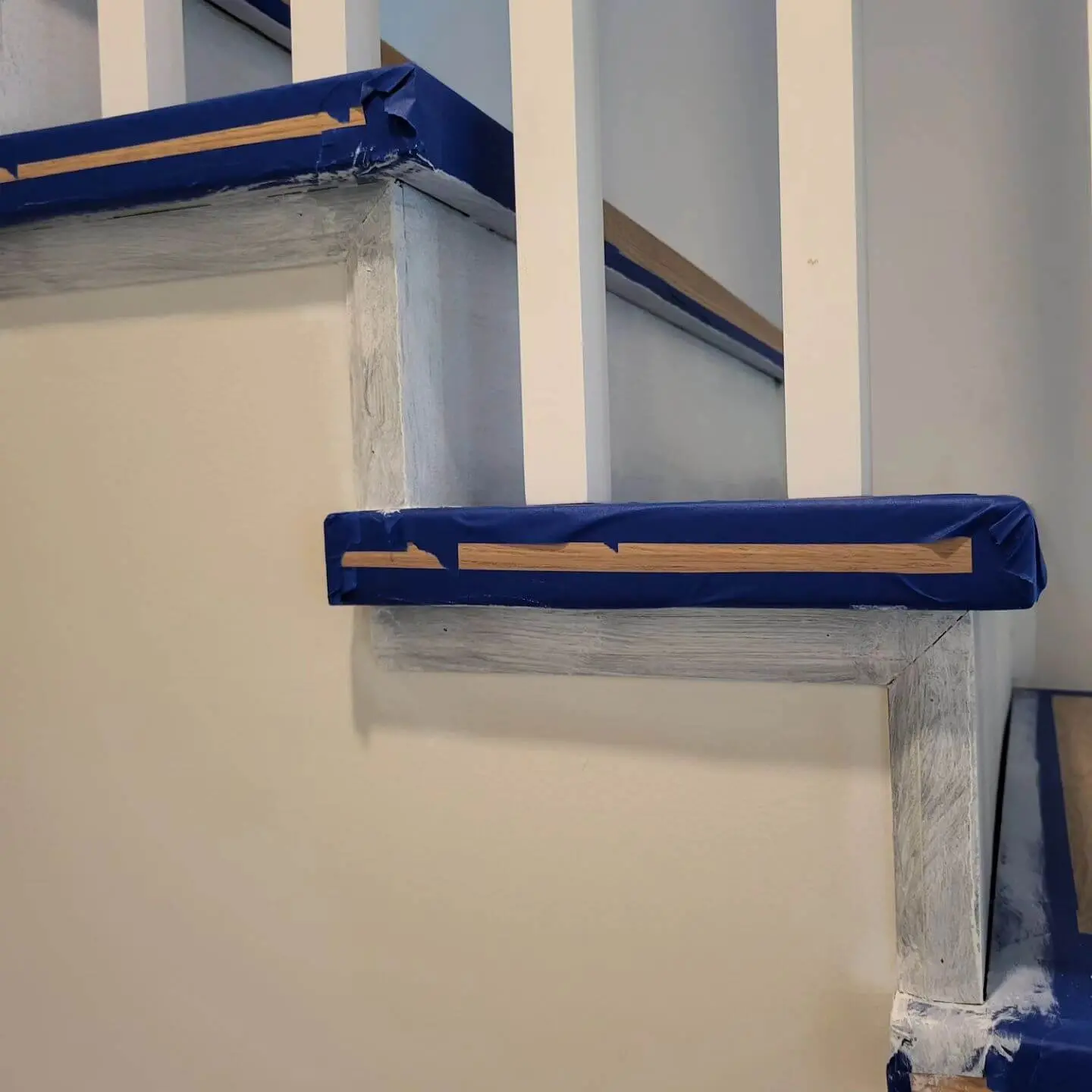 Detailed view of staircase trim and risers covered with blue painter's tape during a painting project.