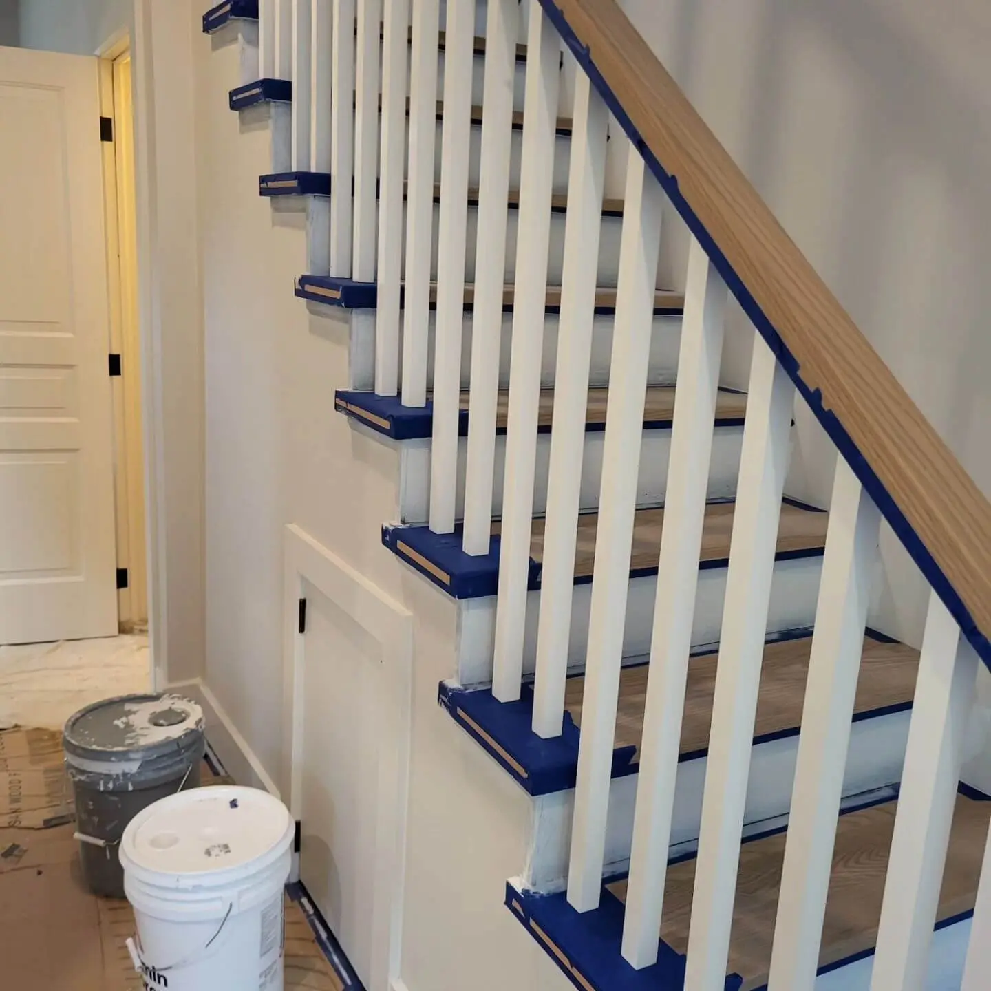 Side view of a staircase with taped balusters, handrails, and steps, alongside painting tools and buckets.